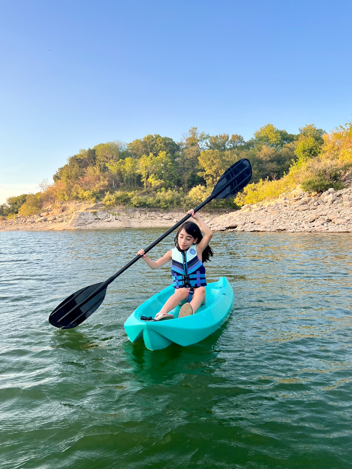 A person is seen paddling a bright aqua kayak on a calm lake, surrounded by gently sloping greenery. The clear blue sky and distant tree-covered hills provide a serene backdrop, while the sun reflects off the water's surface, enhancing the tranquil setting.
