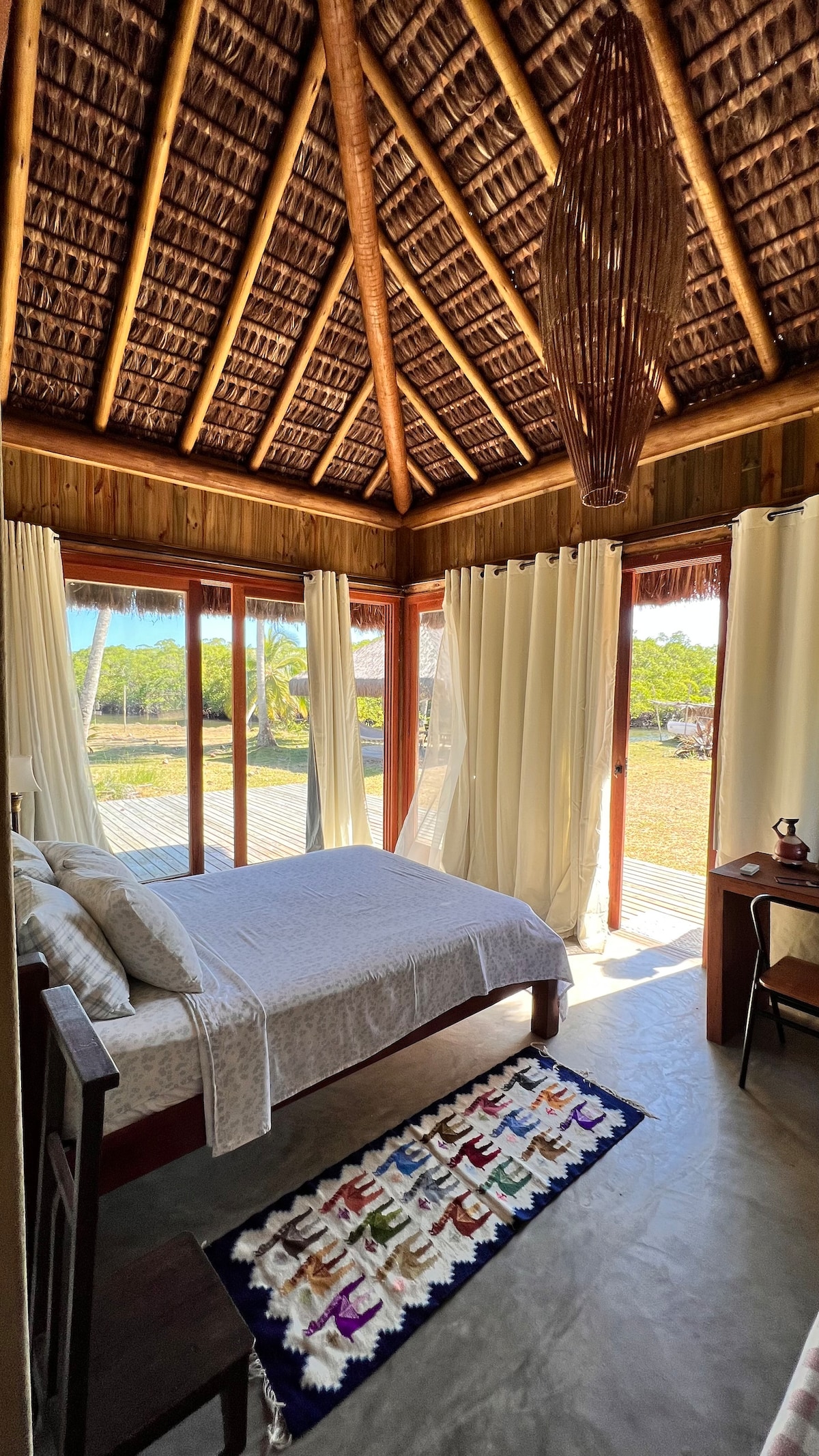 A spacious sleeping area features large glass doors that open to the outdoor view, allowing natural light to fill the room. A decorative rug lies on the floor, and a simple wooden desk and chair complete the cozy, inviting setting.