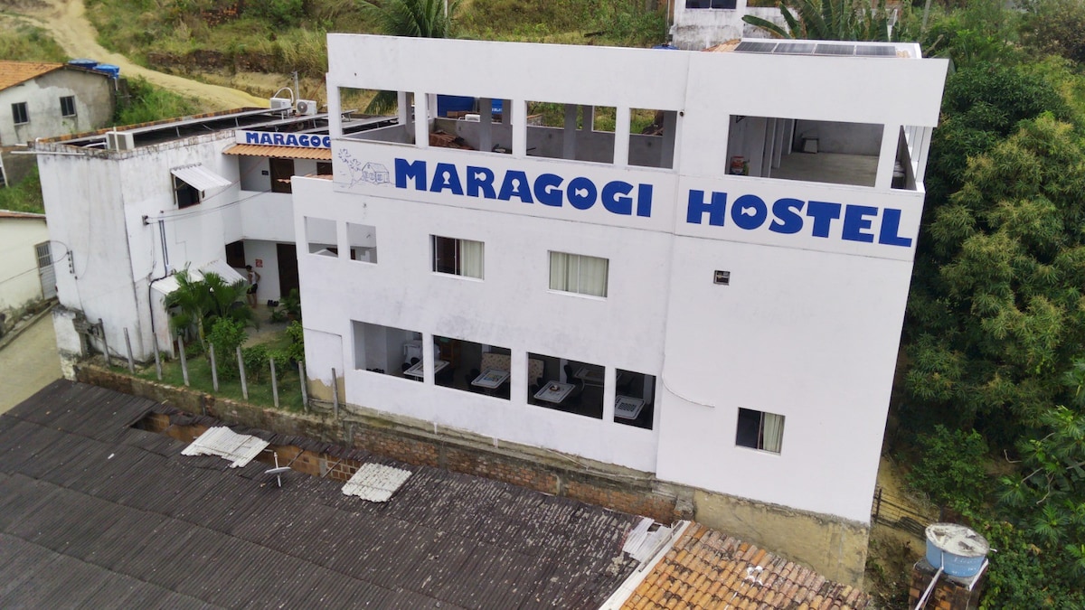 A multi-story white building displays the name 'Maragogi Hostel' in bold blue letters. Several windows are visible, showcasing a balanced arrangement across the facade. Surrounding greenery adds a natural element to the setting, complementing the hostel's structure.