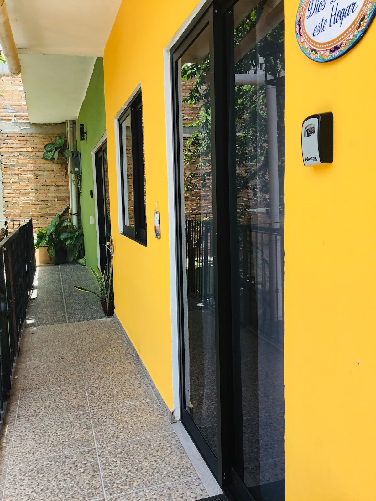 A brightly painted yellow exterior leads to an entryway with glass doors. The corridor features a smooth surface and is adorned with greenery, creating a welcoming path to the nearby apartments. Natural light reflects softly off the polished floor.