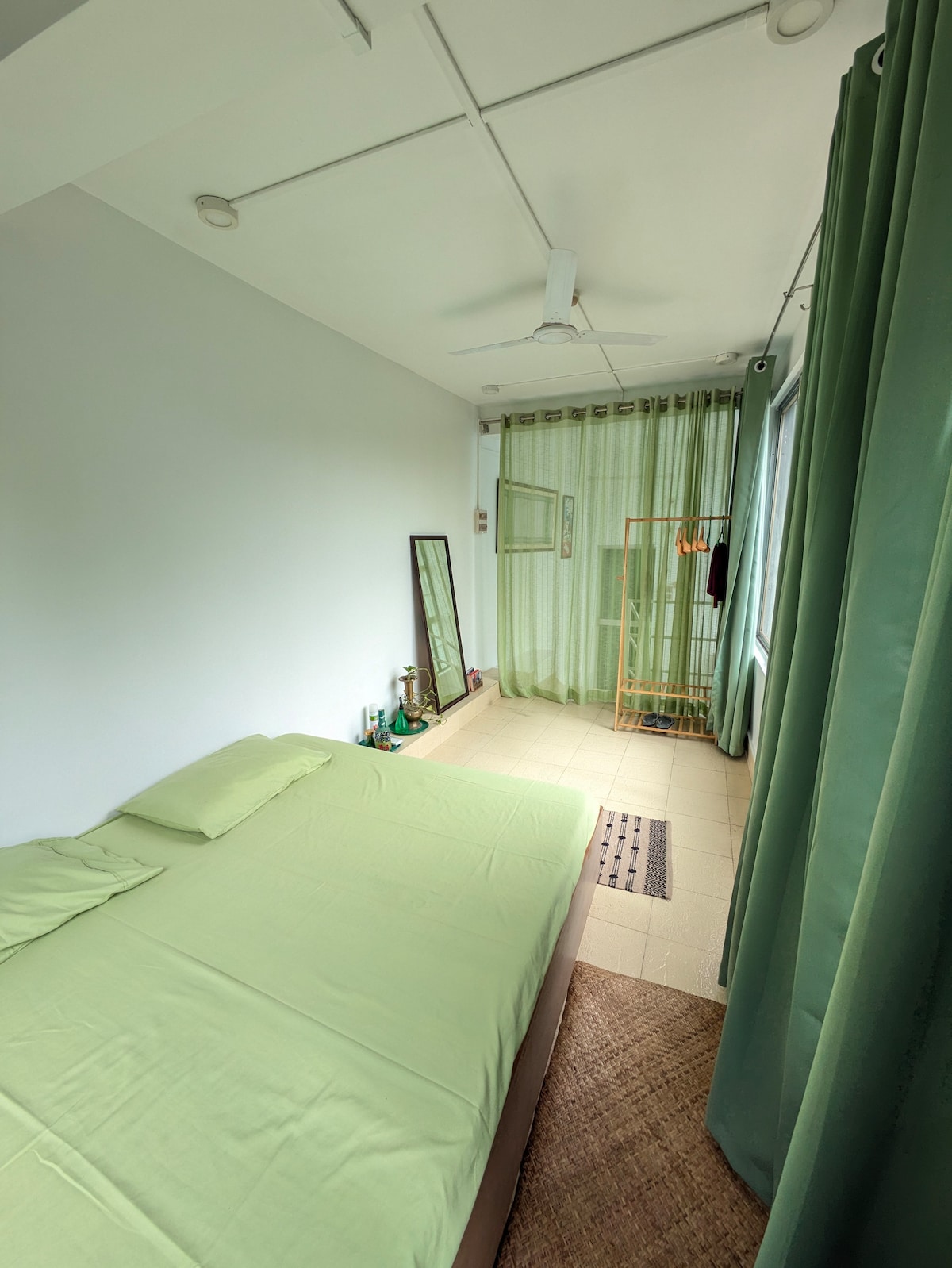 A serene sleeping area is featured with a soft green bedding set atop a spacious bed. Light green curtains frame the window, allowing natural light to fill the room. A mirror and a decorative hanger stand beside the bed, contributing to the uncluttered ambiance.
