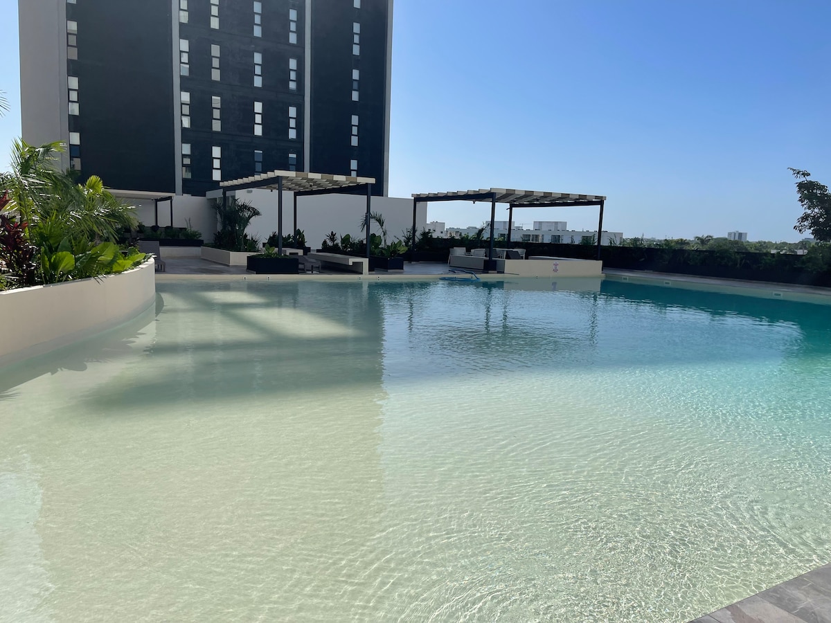 A large, serene swimming pool is shown, with clear turquoise water reflecting the sky. Surrounding the pool, lush greenery and shaded areas create a tranquil environment. The modern building stands in the background against a bright blue sky.