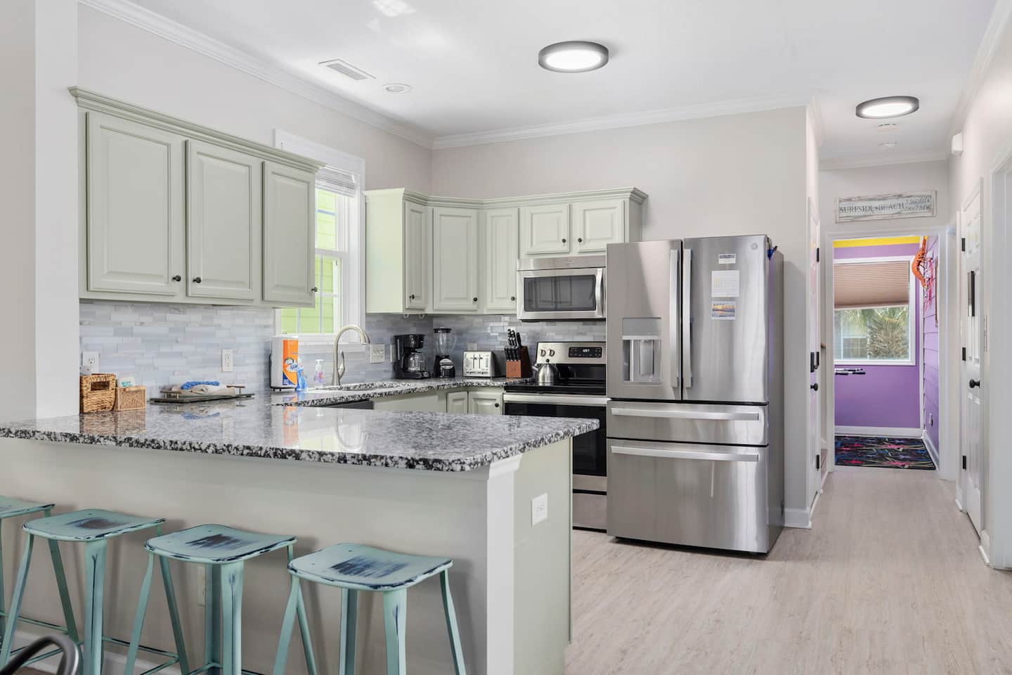 Cook at the granite peninsula with teal bar stools for the sous chefs. Sage green cabinets, stainless appliances, and the arcade room visible down the hall.