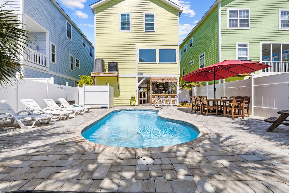Take in the full backyard in one view: pool, tiki bar, dining umbrella, lounge chairs, all of it yours for the week.
