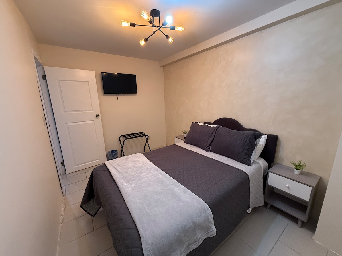 A well-organized bedroom is presented with a spacious bed dressed in dark linens. A soft gray throw is placed at the foot of the bed. A small bedside table holds a lamp, and a wall-mounted television is positioned at eye level. A folding luggage rack is also included.