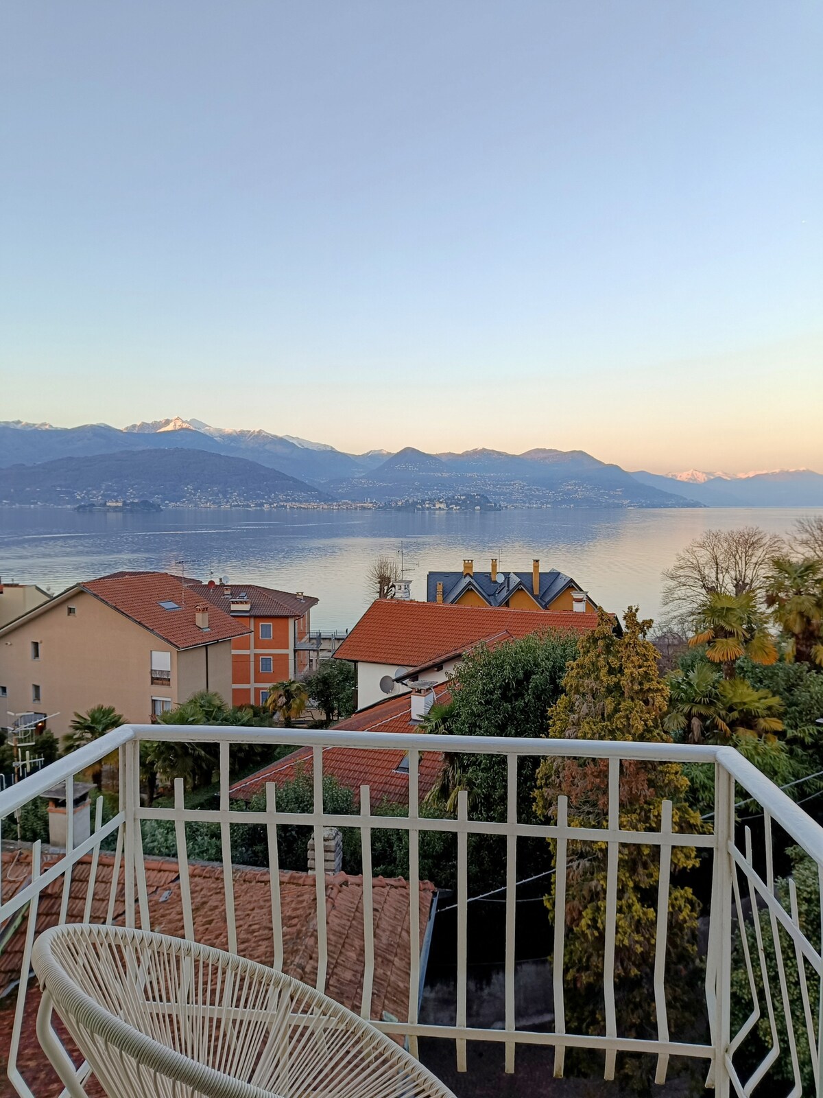 A balcony view showcases a serene landscape, featuring the lake and mountains in the background. Vibrant rooftops of nearby homes blend with lush greenery, while the sun casts a gentle light across the scene, highlighting the natural beauty of the surroundings.