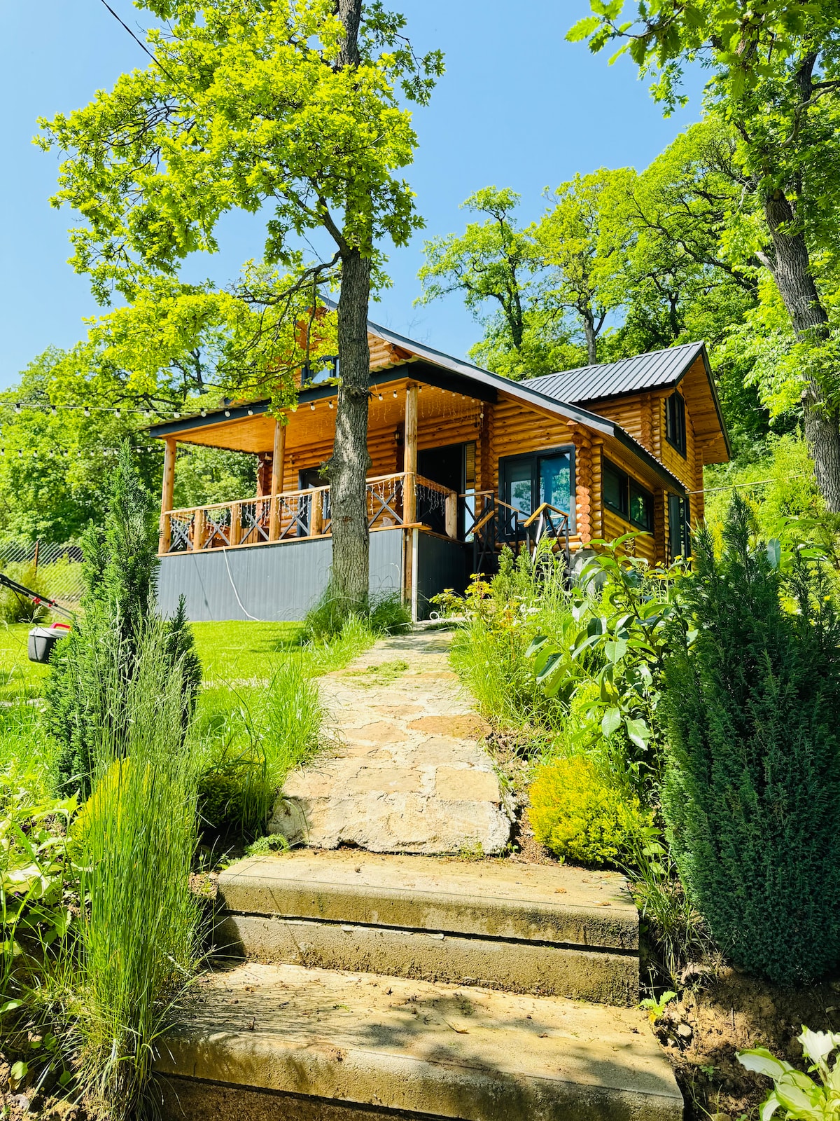 A charming wooden cottage is nestled among greenery, with a wraparound porch and large windows. Stone steps lead through lush plants and grass, enhancing the connection to nature. The setting features a bright sky above, contributing to the tranquil atmosphere of the surrounding landscape.