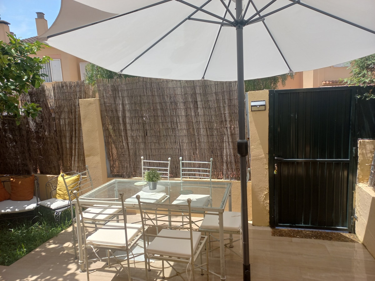 A shaded outdoor seating area features a glass dining table surrounded by white chairs. A large umbrella provides cover, enhancing comfort. Lush greenery frames the space, with a gated entrance visible in the background, creating a private and inviting atmosphere.