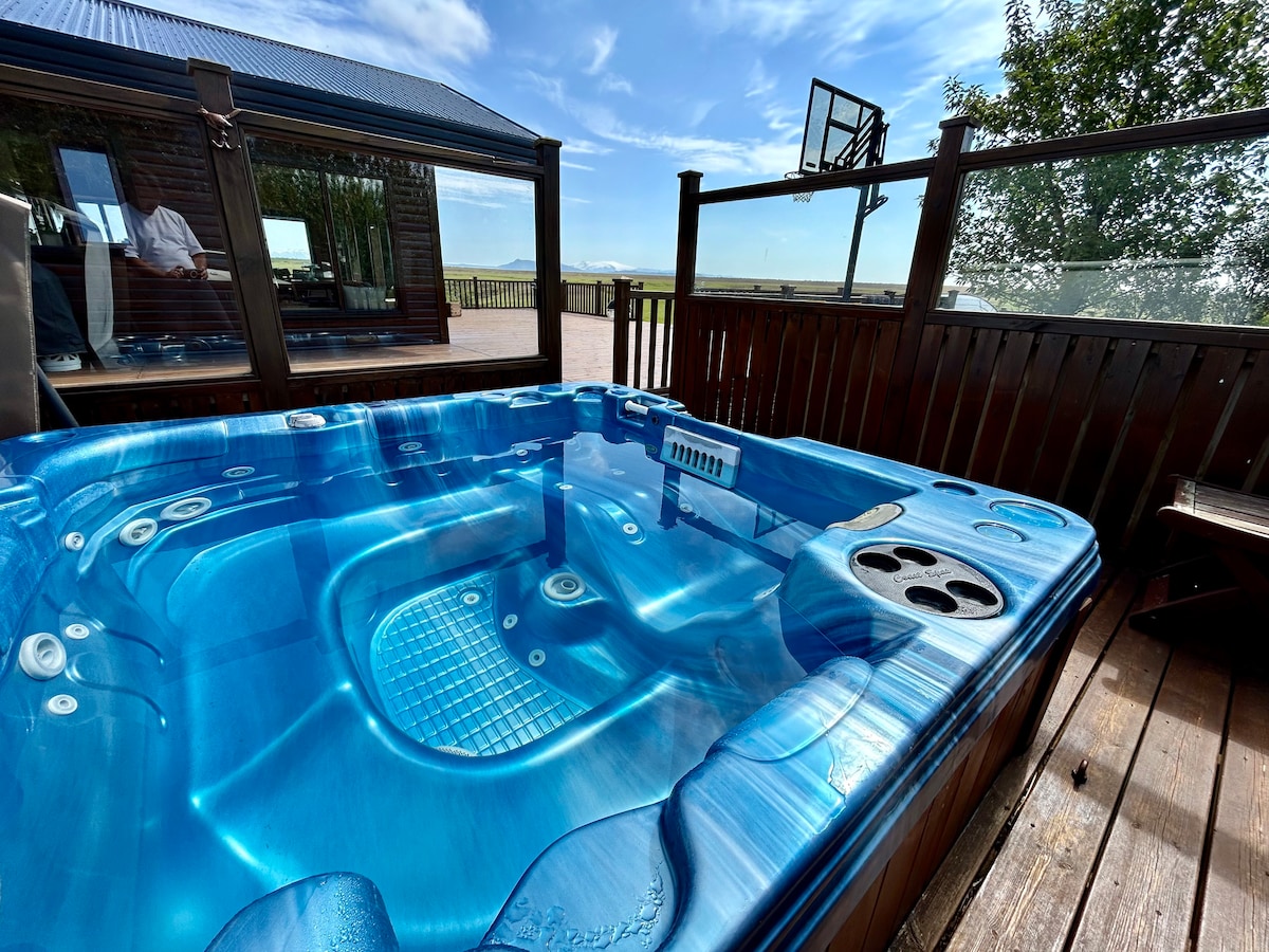 A well-maintained hot tub is featured on a wooden deck, surrounded by glass panels. The water surface reflects the clear blue sky, with a basketball hoop visible in the background. The hot tub includes built-in seating and cup holders for convenience.