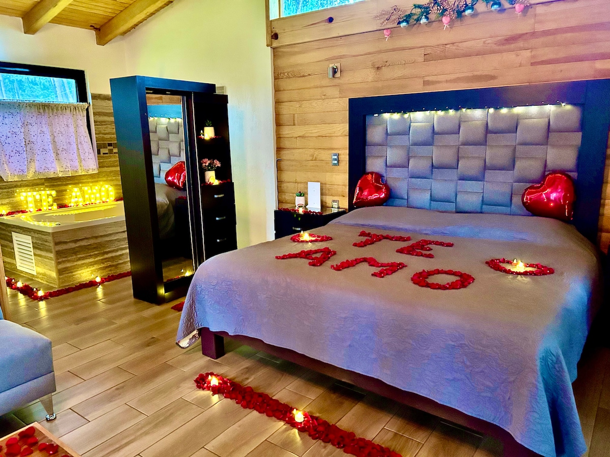A cozy suite is presented with a large bed adorned with rose petals arranged in a heart shape. Soft lighting and candles create a romantic ambiance. A jetted tub is visible in the background, complemented by dark wood accents. A chic cabinet stands nearby, adding to the decor.