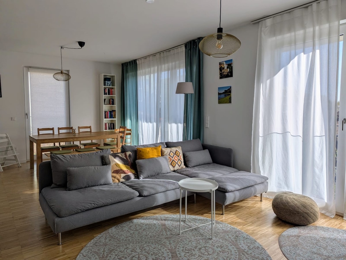 A bright living area includes a spacious gray sofa with decorative cushions, paired with a small white side table. Natural light filters through sheer curtains, highlighting a large dining table and a bookshelf in the background. A cozy rug anchors the space.