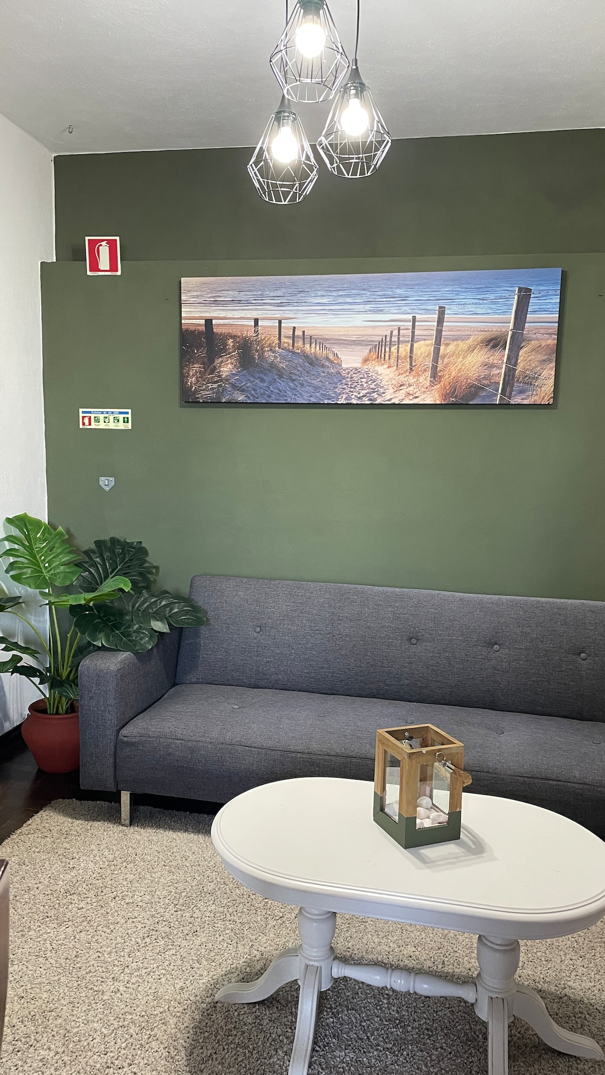 A cozy seating area features a gray sofa against a dark green wall, complemented by a small round table with a decorative lantern. A framed landscape artwork depicting a serene beach scene hangs above the sofa, while a potted plant adds a touch of greenery.