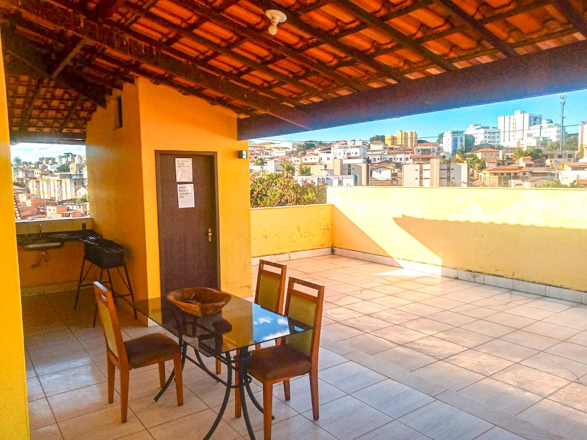 An outdoor terrace is featured with a tiled floor and a wooden ceiling, offering a spacious area for relaxation. A glass-top table with four chairs is arranged near a barbecue grill. The backdrop includes a panoramic view of urban buildings and greenery.