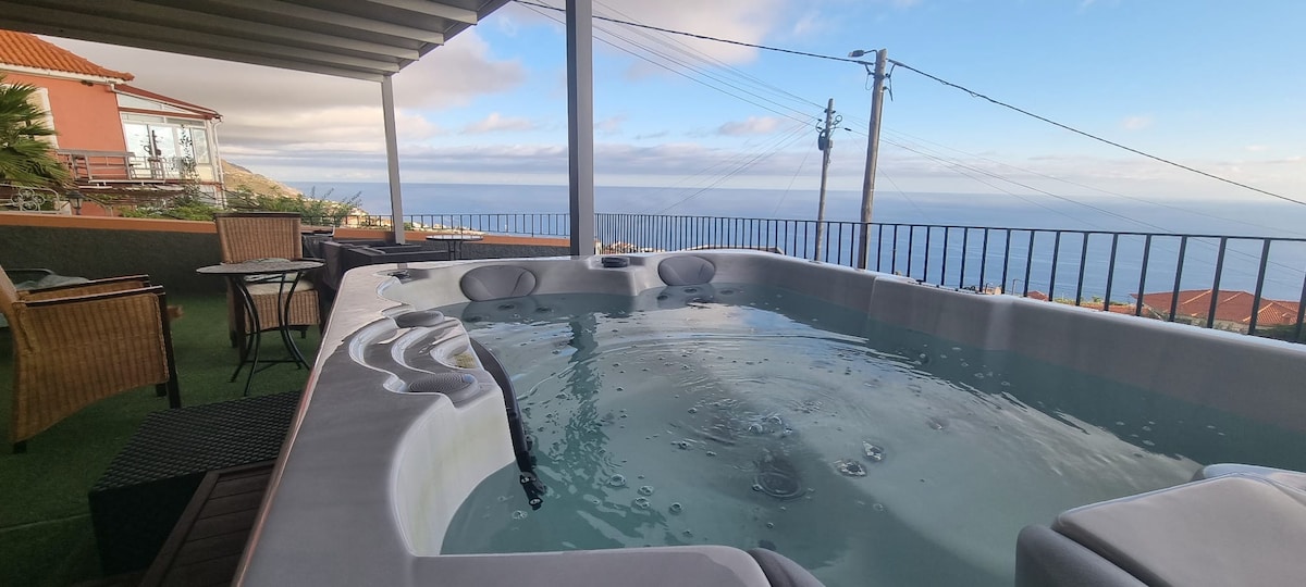 A jacuzzi surrounded by a protective railing is visible, offering a panoramic view of the ocean and distant landscape. A small table and chairs are nearby on a green surface, creating a relaxing outdoor space.