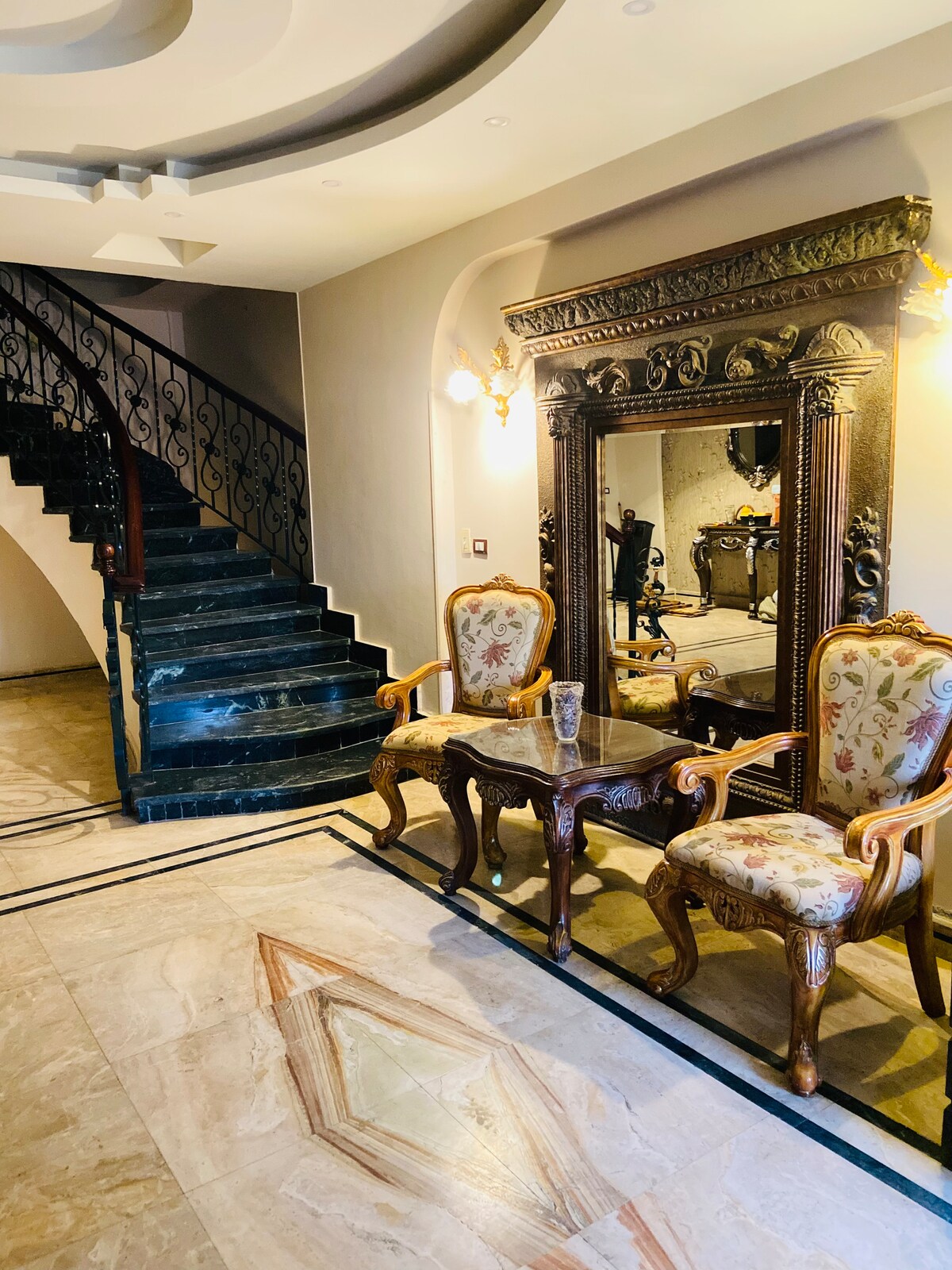A grand entryway features a curved staircase with an ornate railing. Two elaborately designed chairs are positioned near a large, decorative mirror. The marble flooring showcases intricate patterns that enhance the elegant ambiance of the space.