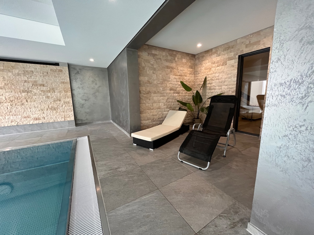 An indoor area features a sleek pool with a metallic edge, surrounded by gray stone walls. A black lounge chair rests beside the pool, complemented by a potted plant nearby and a door leading to another room.