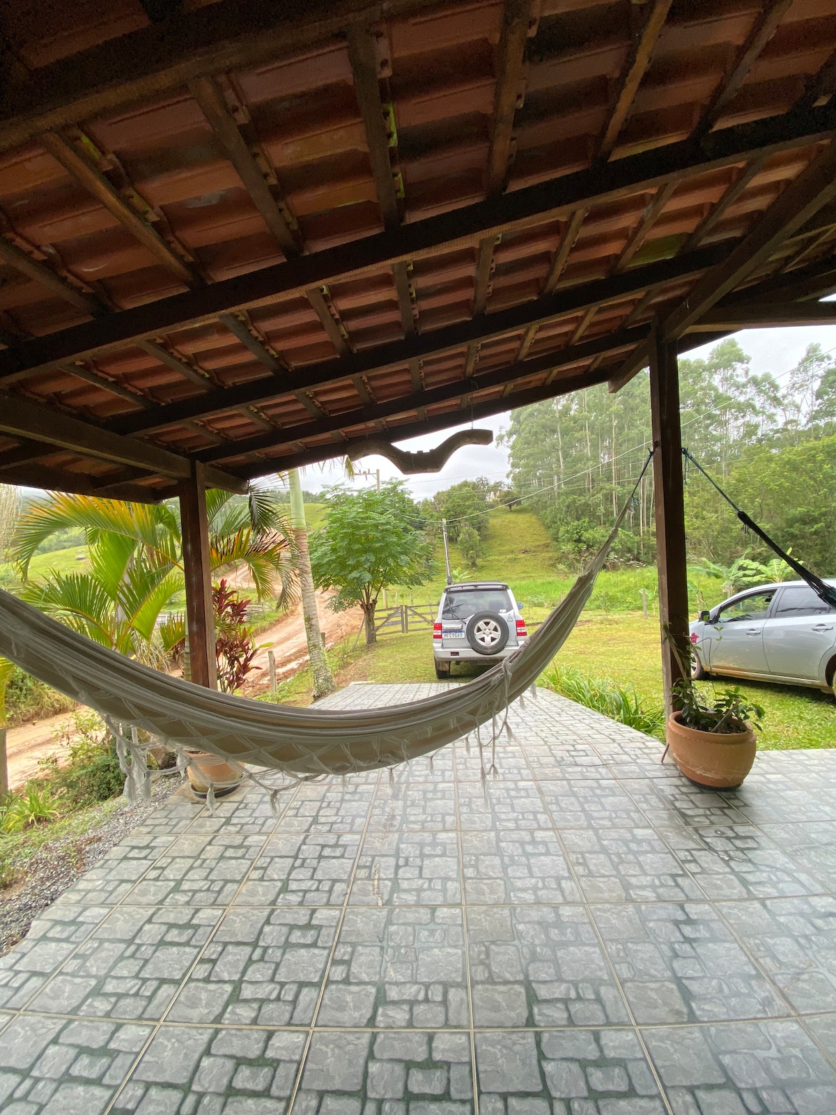 A covered porch is visible, featuring a hammock gently swaying. Potted plants adorn the corners, while the view reveals lush greenery and distant trees. Two vehicles are parked on the gravel path, highlighting the tranquil outdoor setting.