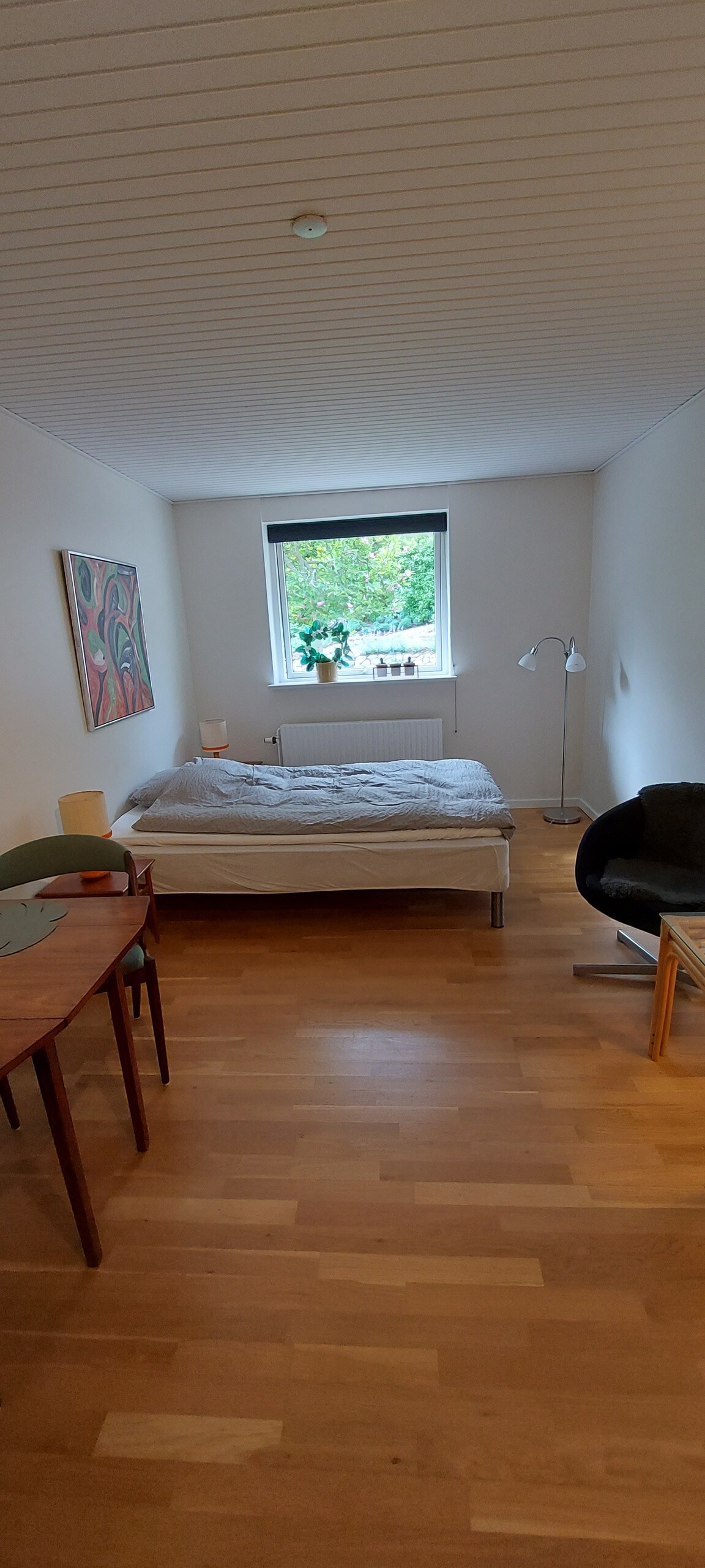 A well-furnished room features a spacious bed and a desk with two chairs. Natural light enters through a window, complemented by a floor lamp nearby. A small area with a cozy chair invites relaxation, while wooden flooring adds warmth to the space.