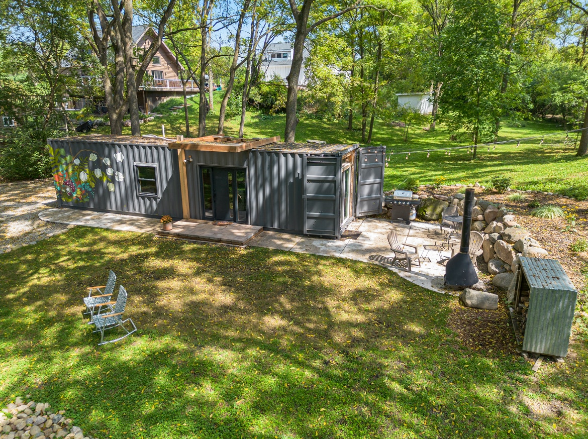 The shipping container tiny house is nestled on a grassy area, surrounded by trees. A private stone patio features mid-century outdoor fireplace and seating options. Sunlight filters through the leaves, creating a serene outdoor setting. Nearby, additional greenery enhances the natural surroundings.