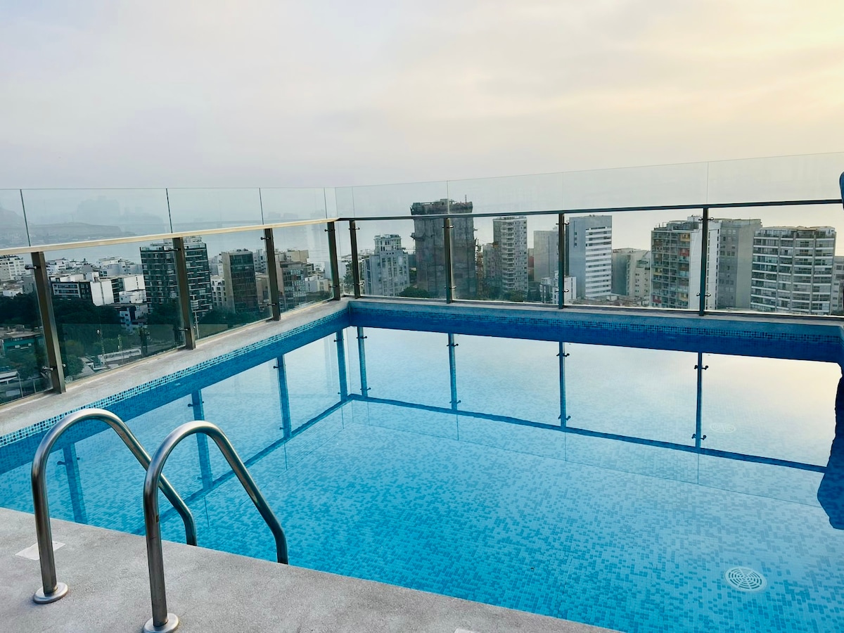 An expansive rooftop pool is framed by a glass railing, offering panoramic views of the city skyline. The water's surface reflects the soft hues of the sky, creating a serene relaxation space for guests. Steps lead into the pool from the surrounding deck.