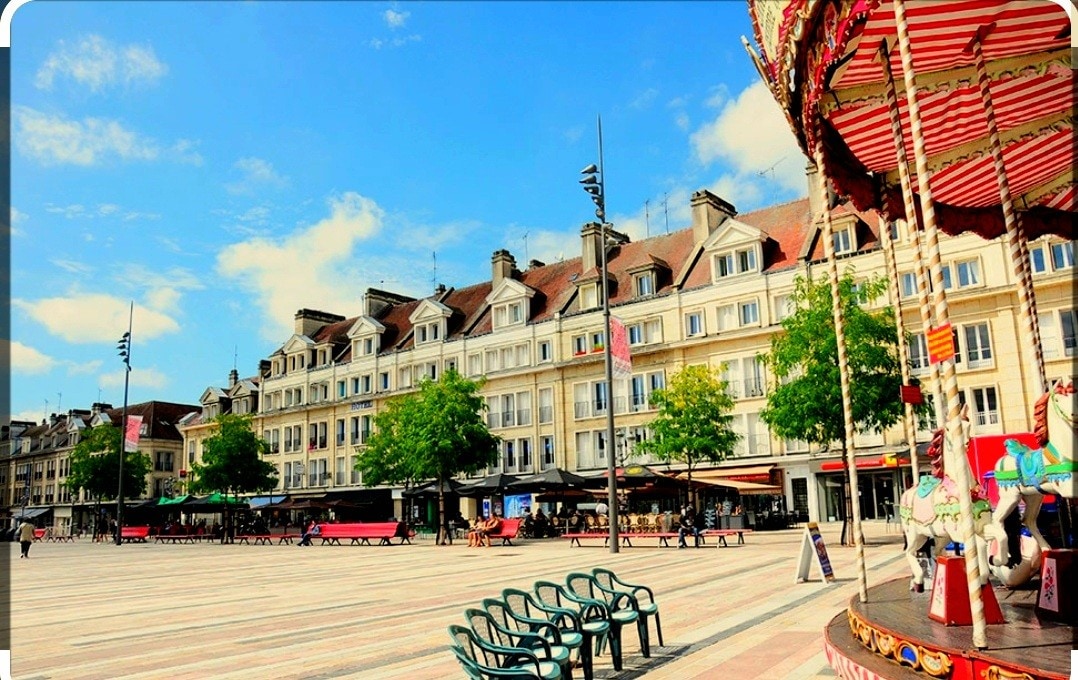 A vibrant town square is featured, showcasing a charming carousel to one side. Tree-lined walkways are visible, accompanied by rows of buildings characterized by classic architecture. Public seating areas with green chairs provide a welcoming atmosphere for visitors, under a clear blue sky.