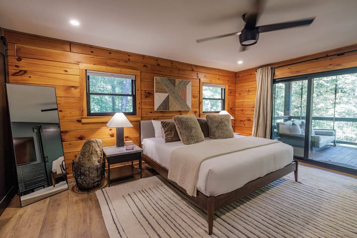 The king-sized primary suite features a West Elm bed, faux fur accents, and two doors opening directly to the covered porch — wake up to the sound of the Ellijay River.