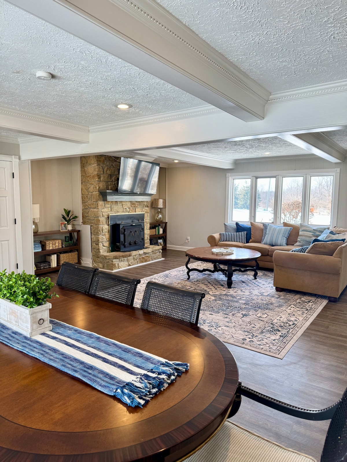 A spacious living area features a comfortable seating arrangement with a neutral-toned couch and a patterned area rug. A stone fireplace serves as a focal point, while large windows allow natural light to illuminate the space. A dining table with decorative accents is visible in the foreground.