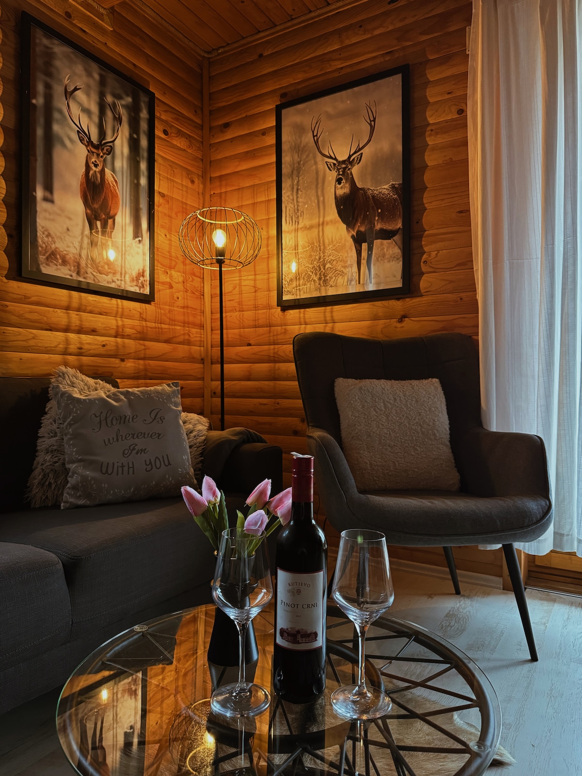 A cozy sitting area features wooden walls, two framed deer prints, and a warm light fixture. A round glass coffee table holds a bottle of wine and two wine glasses, while a vase with tulips adds a touch of color to the space.