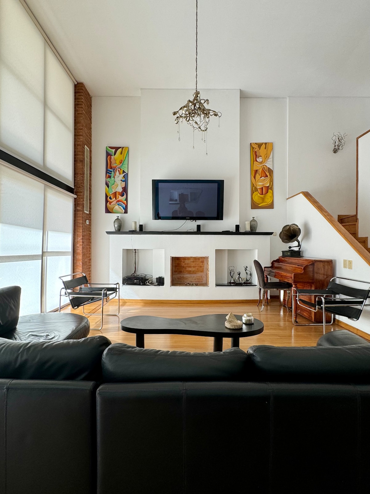 The living area features a modern design with a large black sectional sofa facing a wall-mounted television. Colorful art pieces adorn the walls, while a wooden mantel supports decorative items. A wooden staircase leads to the upper level, providing access to additional spaces.