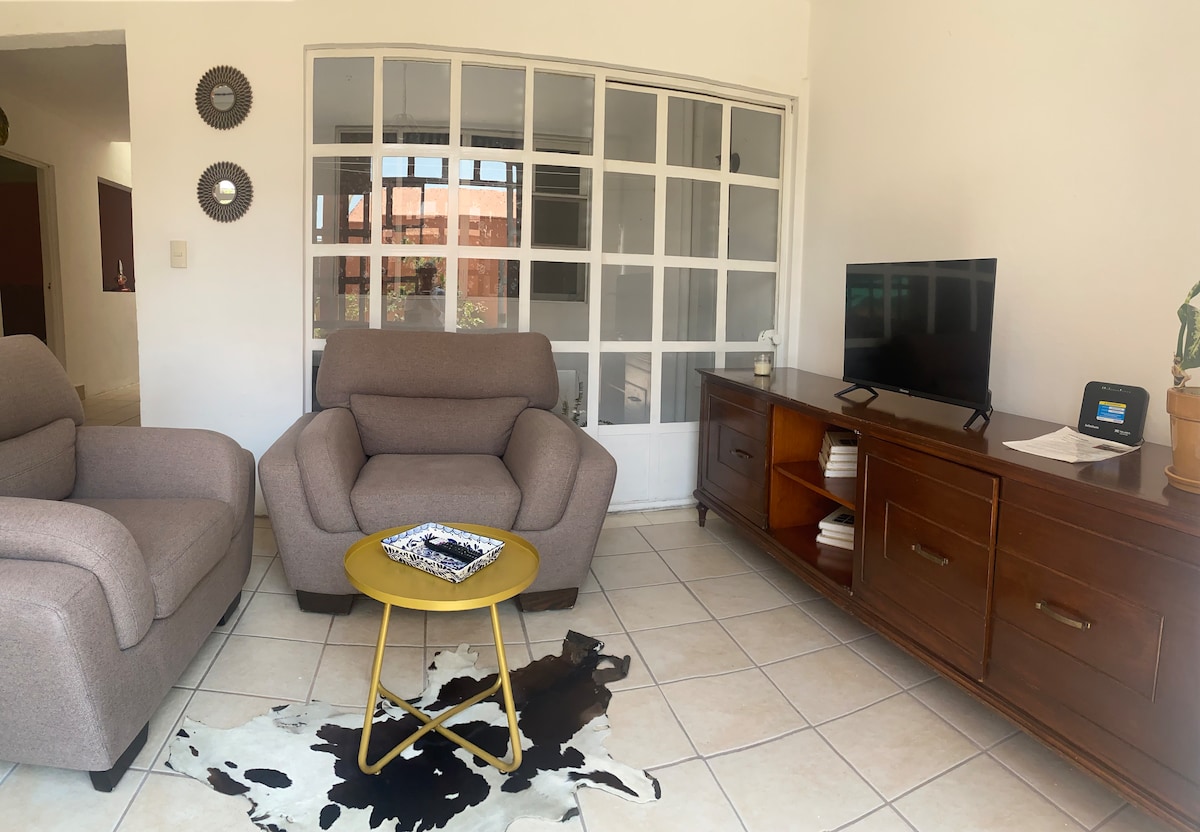 A cozy living area features two comfortable armchairs positioned around a small round table, with a television set placed on a wooden TV stand. Natural light is let in through large glass windows, creating a bright and welcoming space.
