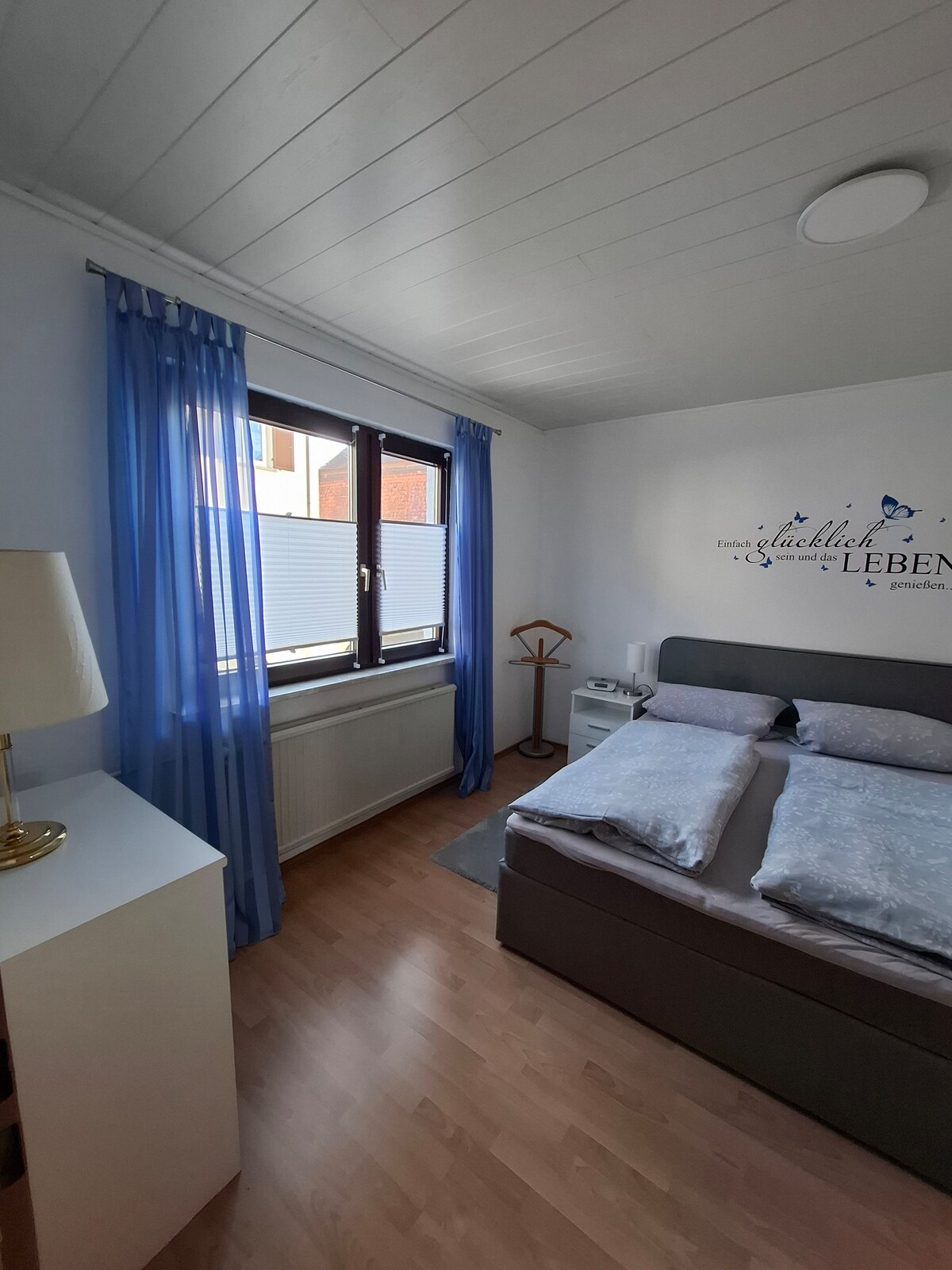 The bedroom features a spacious double bed with light bedding. Tall windows dressed in sheer blue curtains allow natural light to enter, while a bedside table and a lamp provide additional lighting. A small wardrobe is also visible, enhancing the room's storage options.