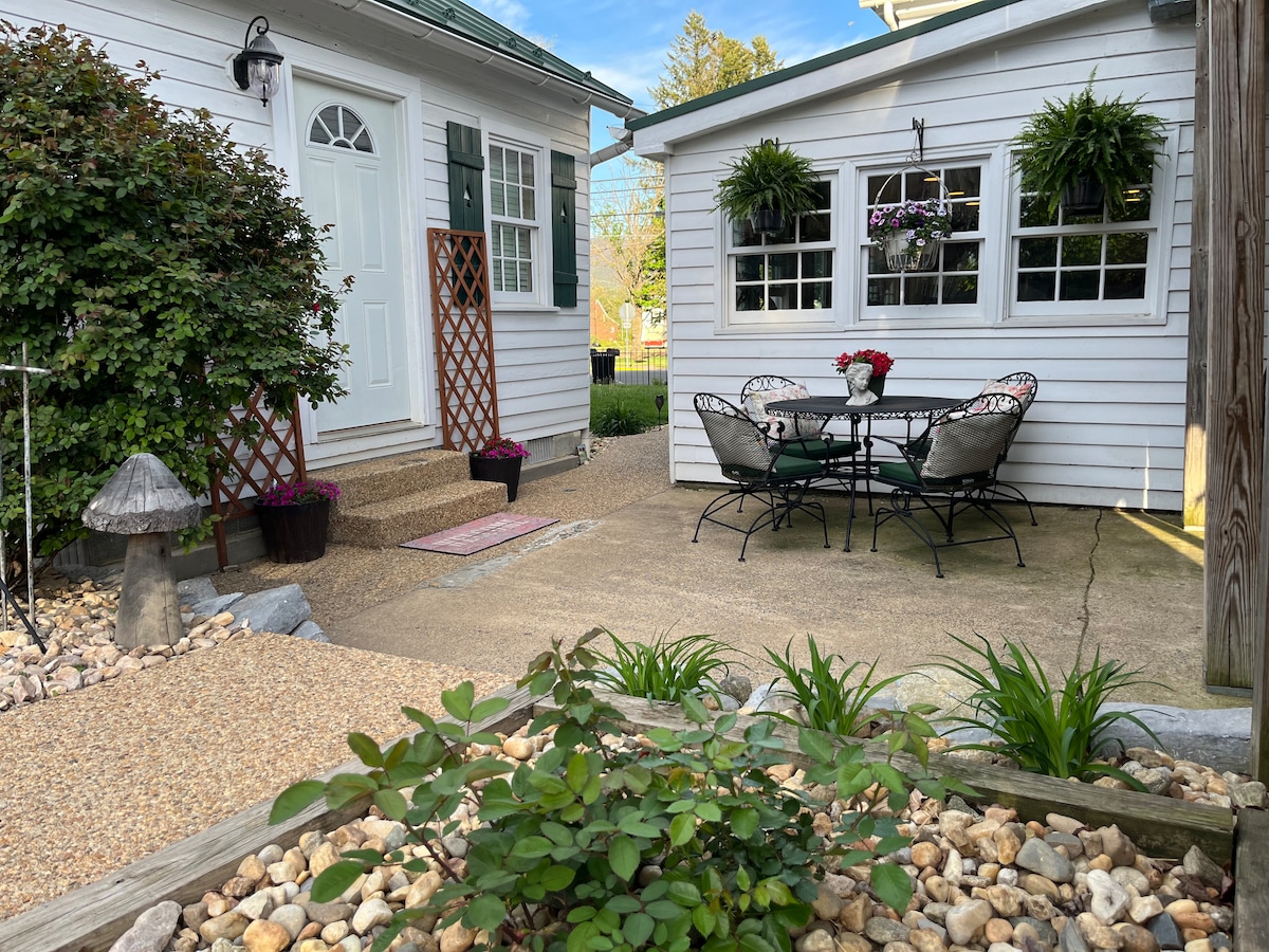 A charming outdoor seating area is shown, featuring a small round table surrounded by four metal chairs. Potted plants and hanging baskets add greenery, while a stone pathway leads to the entrance of the cottage. A decorative mushroom sits among the pebbles in the foreground.