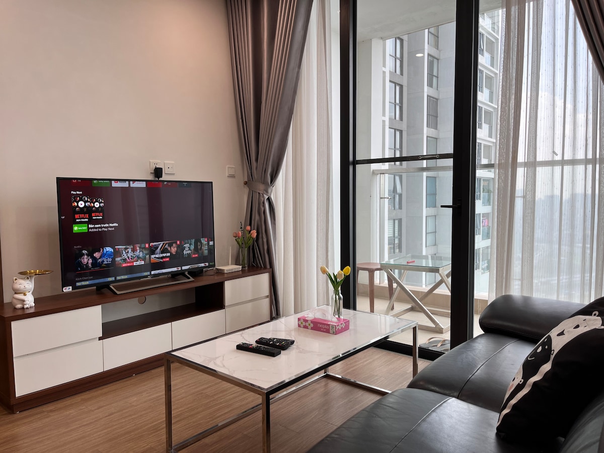 A modern living room features a large flat-screen TV mounted on a white entertainment unit. A sleek coffee table sits in front of a black sofa. Floor-to-ceiling windows allow natural light to fill the space, with sheer curtains softly framing the view.