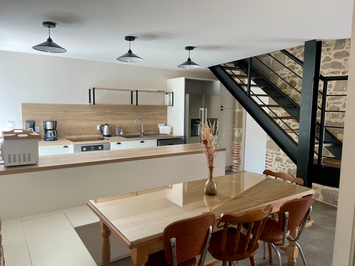 The open kitchen features a modern design with a central island and ample countertop space. Kitchen appliances are neatly arranged, including a coffee maker and kettle. A dining table with wooden chairs is positioned nearby, complemented by exposed stone walls and a staircase leading to the upper level.