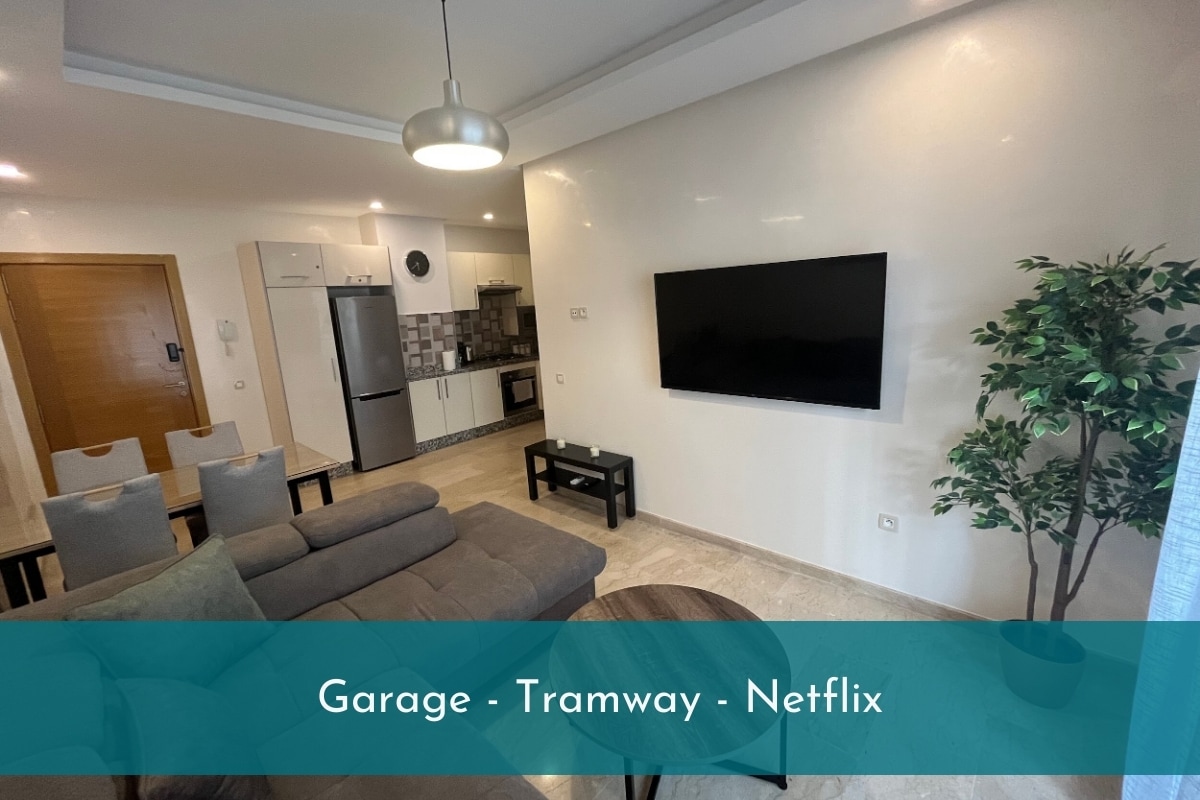 A cozy living area features a spacious sofa and a coffee table, with a large wall-mounted TV displaying Netflix. The open kitchen is visible, equipped with modern appliances, and a small dining area is present. A potted plant adds a touch of greenery.