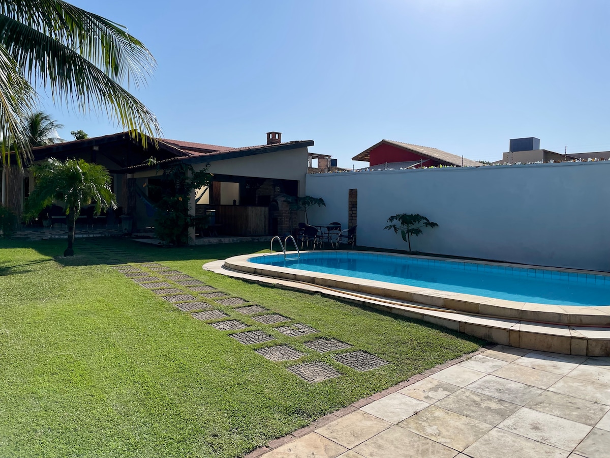 A spacious outdoor area features a clear blue pool surrounded by a manicured lawn. Sunlight reflects off the water as palm trees and a covered patio create a relaxed atmosphere. A pathway of stone tiles leads from the pool to the house in the background.