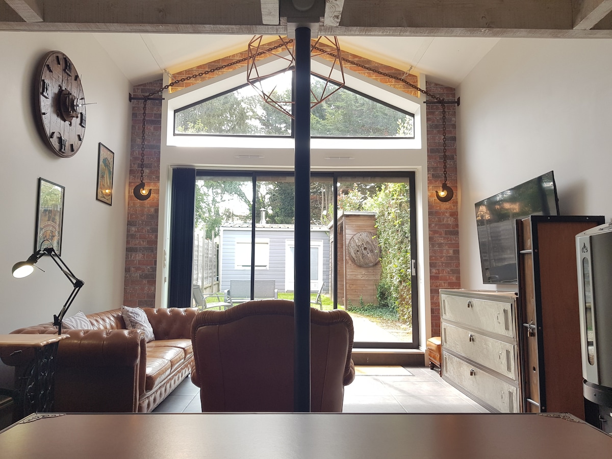 The living area is characterized by high ceilings and large glass doors that open to the garden. A comfortable leather sofa is positioned alongside a wooden cabinet and wall-mounted television. The space features exposed brick walls, decorative wall art, and warm lighting, creating an inviting atmosphere.