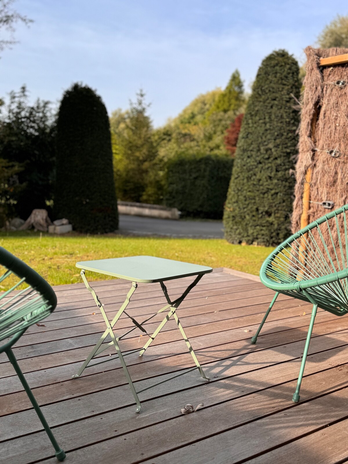 A wooden deck features two green chairs positioned beside a small folding table. In the background, well-maintained evergreen trees provide a lush and private setting, complemented by natural green surroundings and a clear sky.