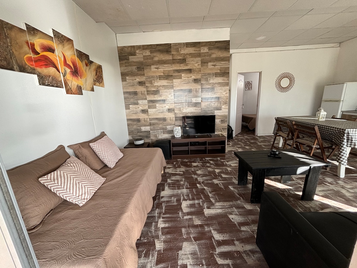 A living area is presented with a neutral color palette, featuring a comfortable sofa adorned with patterned cushions. A coffee table is centrally positioned in the space, complemented by a wall-mounted television. Artwork with floral designs decorates the wall, enhancing the ambiance.