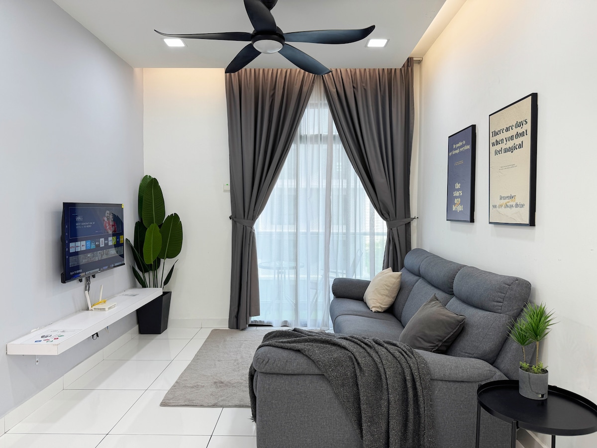 A comfortable living area features a spacious gray sofa adorned with decorative pillows. A large ceiling fan hangs above, and sleek curtains frame the window, allowing natural light to enter. A flat-screen TV is mounted on a minimalist white wall shelf beside a green potted plant.
