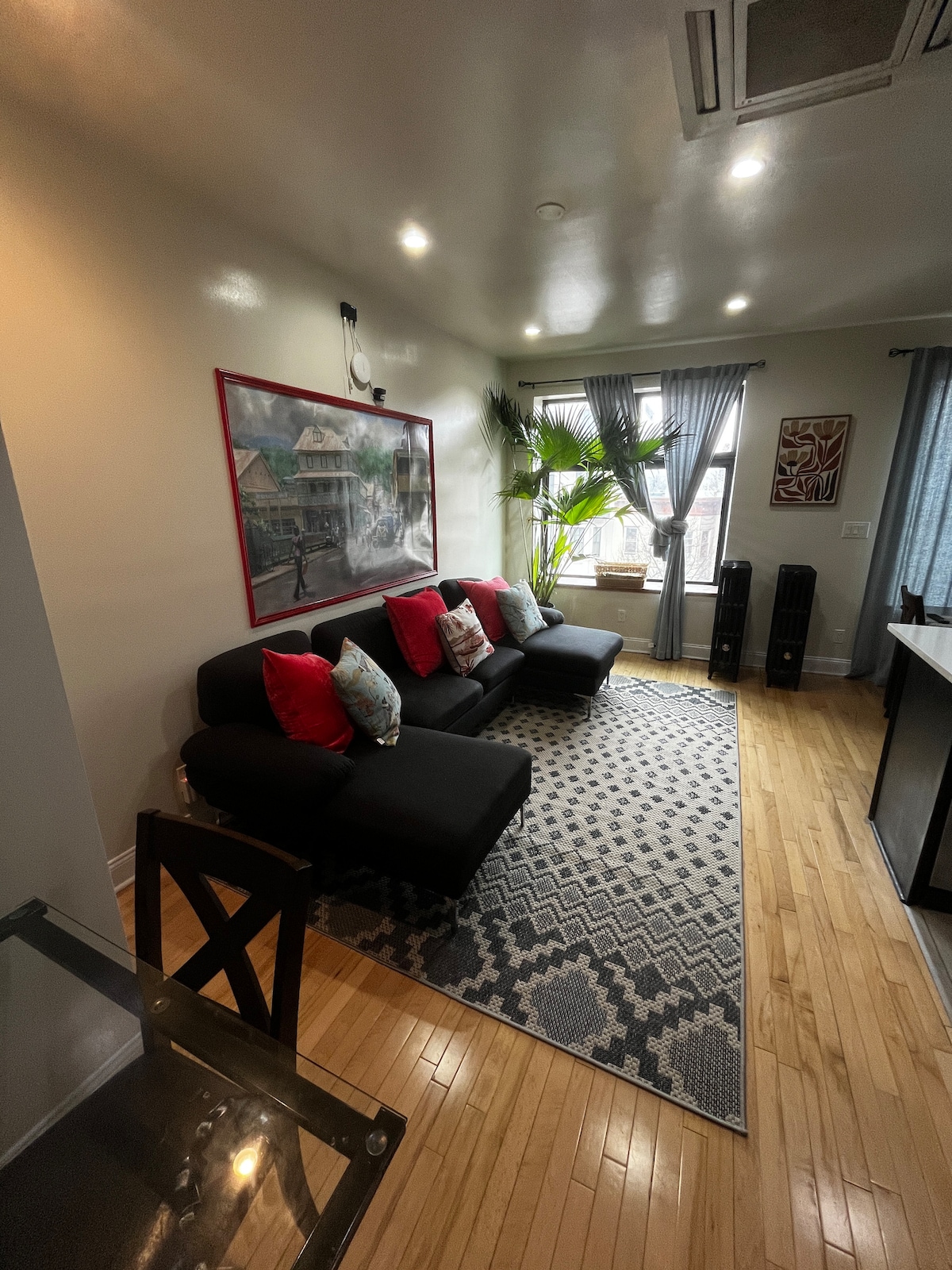 A bright living area features a modern sofa adorned with colorful cushions. A large rug with geometric patterns anchors the space, while a plant adds a touch of greenery. Natural light streams in through window treatments, enhancing the warm ambiance of the room.