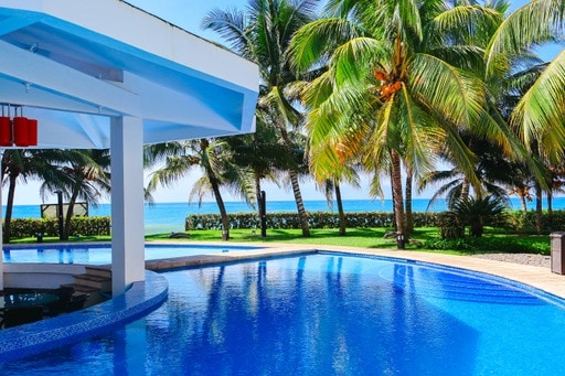 A bright swimming pool glistens under sunlight, surrounded by lush palm trees. The tranquil ocean is visible in the background, complemented by well-maintained green landscaping. The area is designed for relaxation and enjoying the warm outdoor atmosphere.