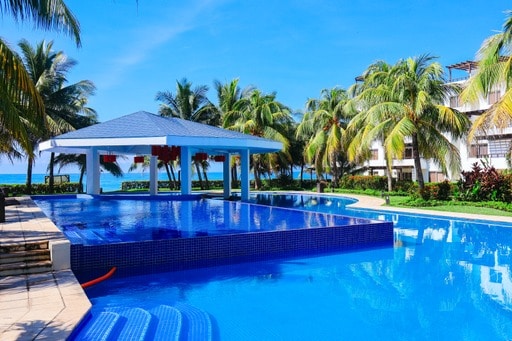 An expansive swimming pool reflects the clear blue sky, surrounded by lush palm trees. A shaded gazebo is positioned nearby, offering a relaxing gathering space. The vibrant colors of the pool tiles contrast with the greenery, creating a calm outdoor setting steps away from the beach.