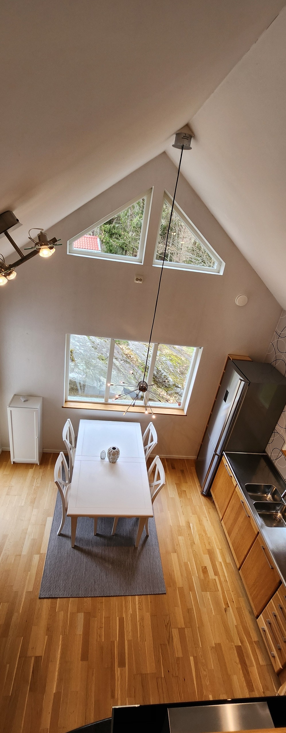 An overhead view of a modern kitchen showcases a spacious dining table surrounded by four white chairs. A large window lets in natural light, highlighting the wood flooring. Stylish lighting hangs above, and sleek kitchen appliances are positioned along the side.
