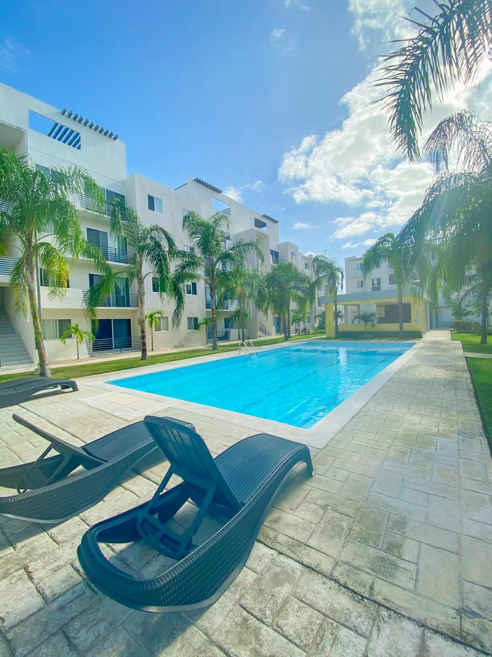 A clear view of a spacious outdoor pool is presented, bordered by lounge chairs and flanked by lush palm trees. Sunlight brightens the area, creating a relaxing ambiance around the well-maintained pool and surrounding lawn.