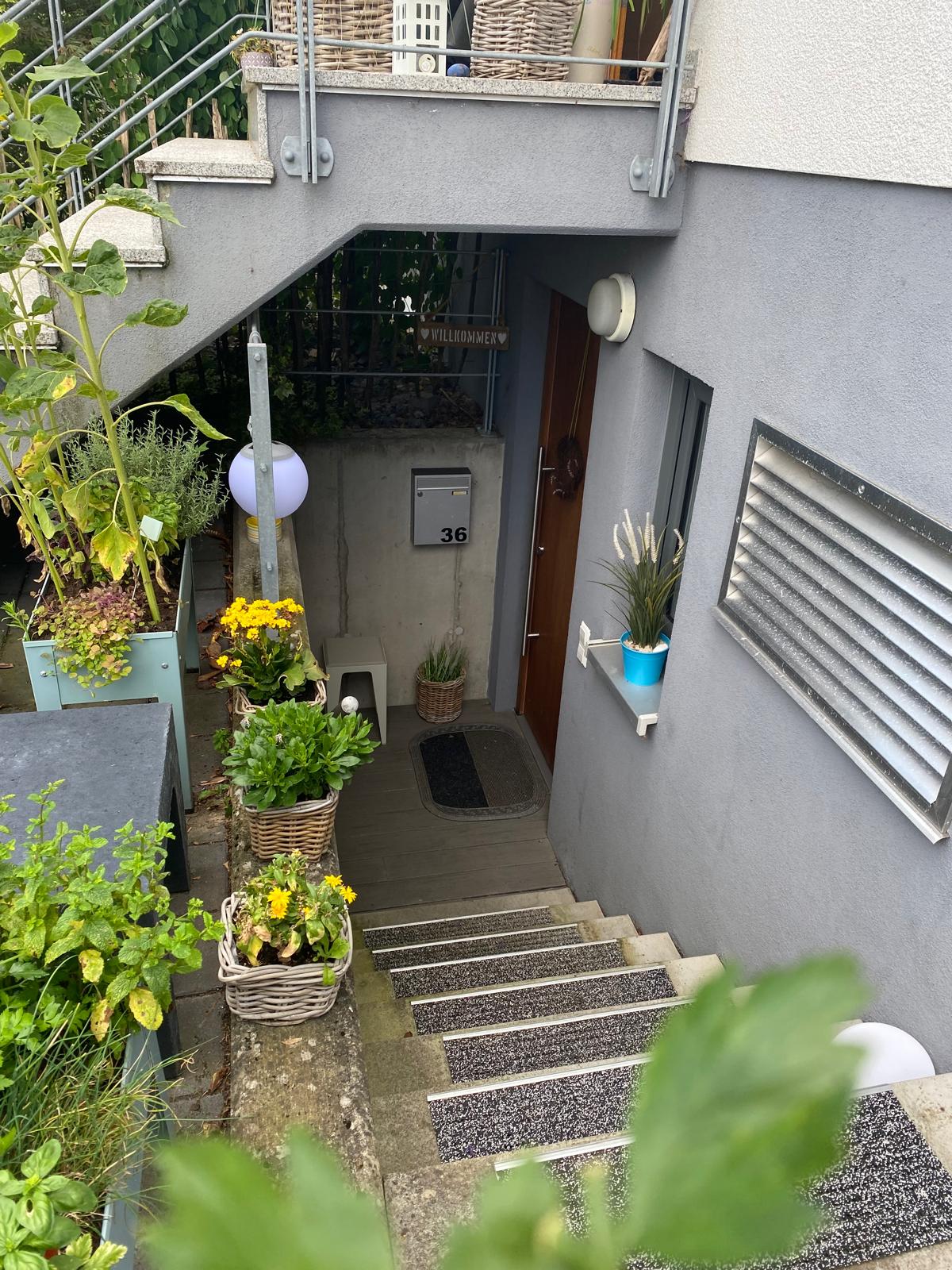 A staircase leads to the entrance of the apartment, flanked by potted plants and greenery. The wall beside the door displays a house number. Soft lighting comes from wall-mounted fixtures, enhancing the welcoming atmosphere of the entrance.