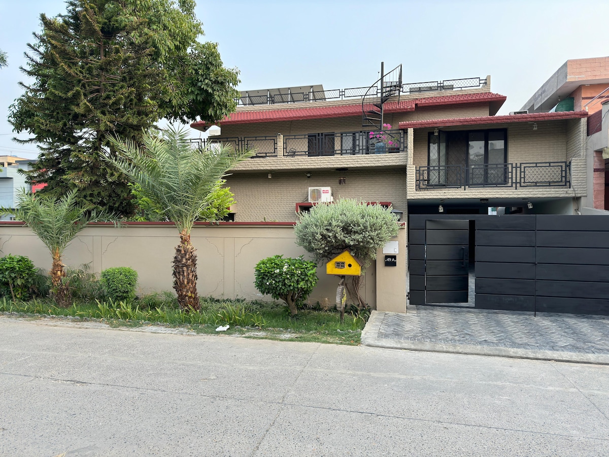A well-maintained exterior of a two-story house is presented, featuring a combination of stone and brickwork. Lush greenery, including palm trees and hedges, is positioned beside a black gate. A pathway leads to the entrance, which is framed by a vibrant wall of flowering plants.