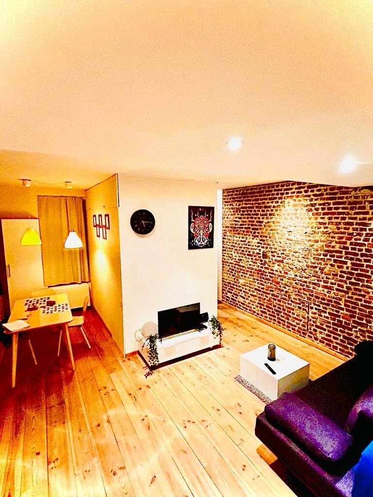 A cozy living area features warm wooden flooring, exposed brick wall, and a sleek coffee table. A comfortable sofa is positioned near a television, with ambient lighting provided by two pendant lamps. A dining table for four is visible, enhancing the functional space.