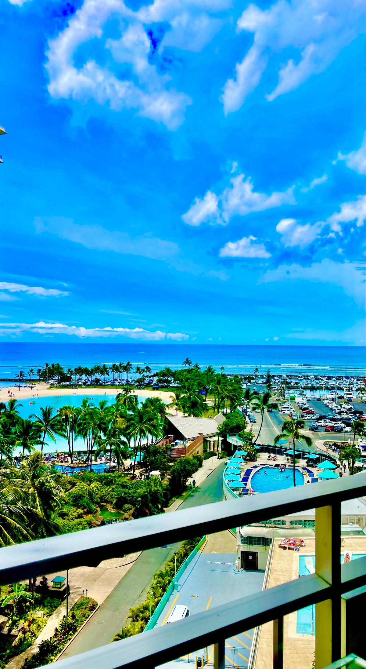 A panoramic view captures the sparkling ocean and the stunning coastline beneath a bright blue sky. Lush palm trees frame the scene, while vibrant pools and a marina filled with boats are visible below, emphasizing the tropical resort atmosphere.