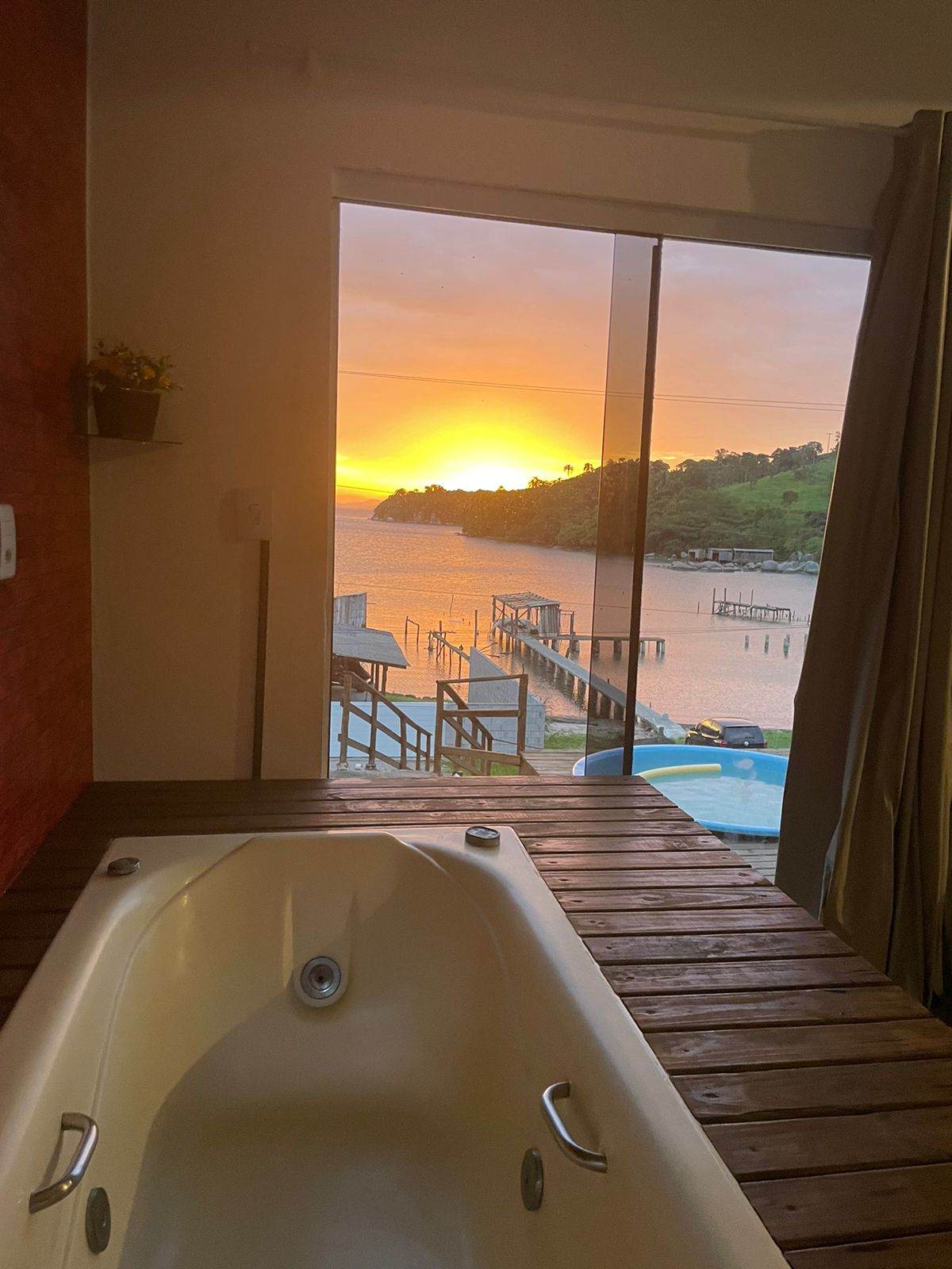 A modern bathroom features a spacious bathtub positioned near a large window. Scenic views of the sunset over a tranquil lagoon and surrounding hills are visible, enhancing the serene ambiance of the space.